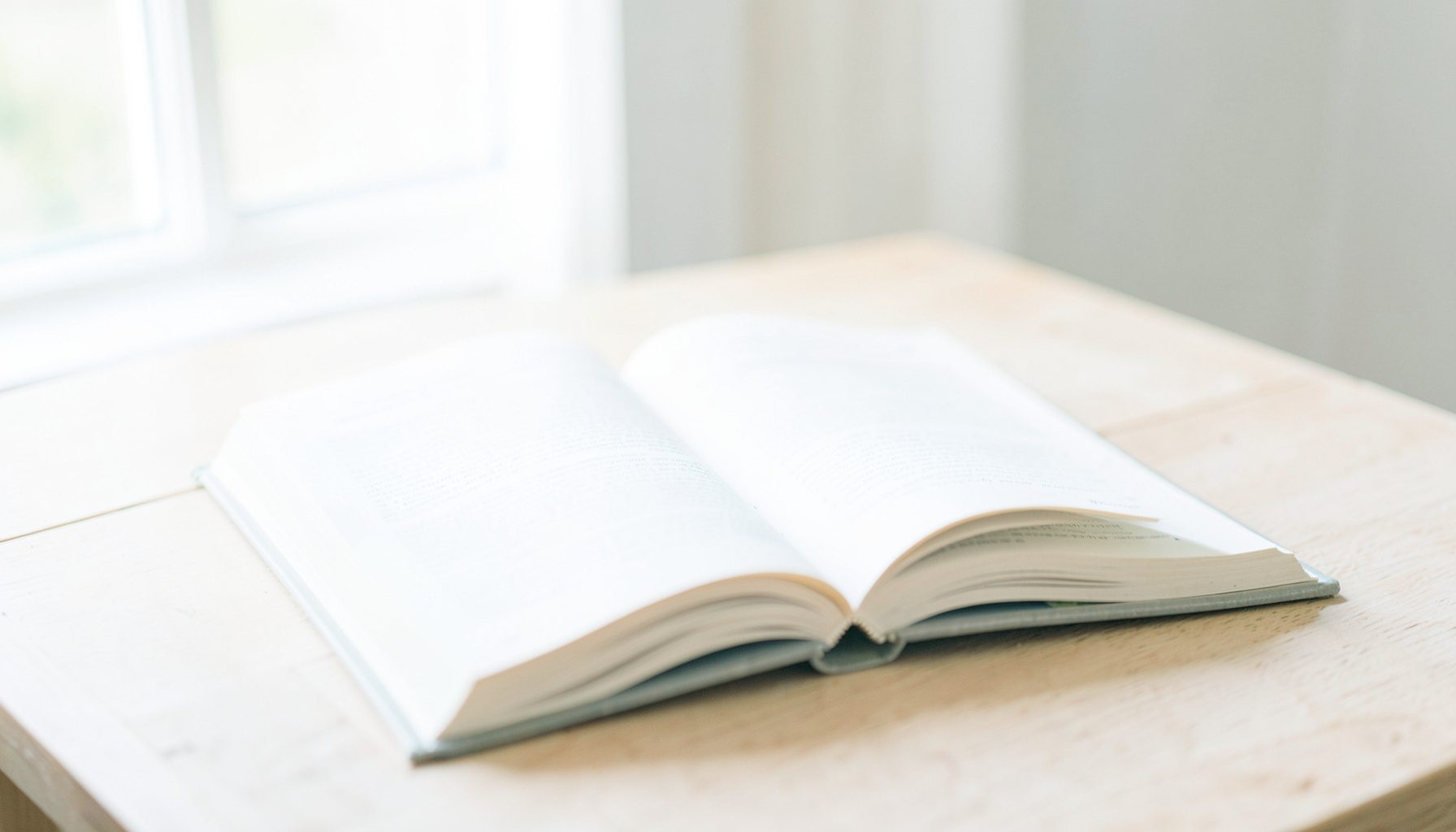 A bright, airy photograph of an open book on a light wooden table near a window, with soft natural daylight and pale cream tones