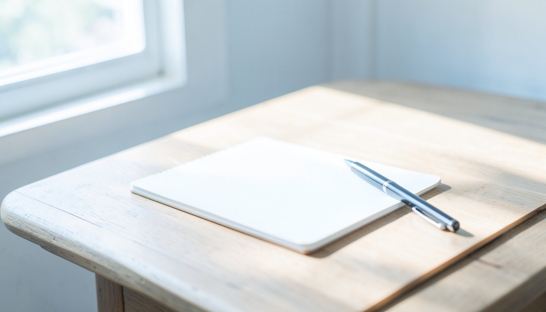 A bright, airy photograph of a wooden writing desk near a sunlit window, with a blank notebook, a pen, and soft natural light casting gentle shadows across the surface.